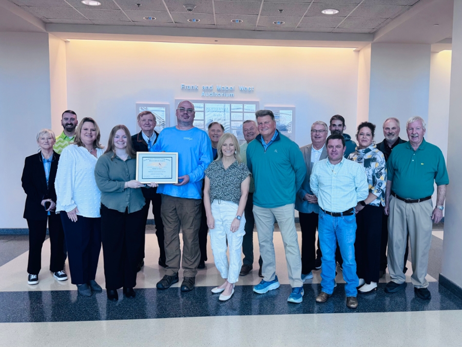 A group of 15 people stands in a well-lit hallway, posing for a photo. A man in a blue shirt and a woman in a green top hold a framed certificate together, smiling. The group, dressed in business casual attire, stands in front of a sign reading 'Frank and Mabel West Auditorium.