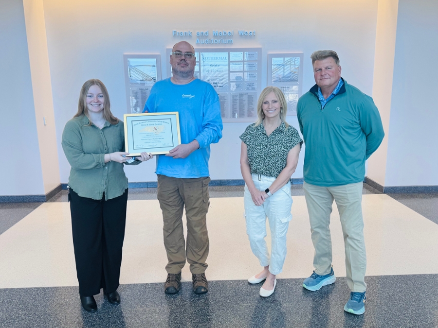 Four people stand in a well-lit hallway, posing for a photo. The person on the left, a woman in a green blouse, holds a framed certificate alongside a man in a blue t-shirt and brown pants. Two other individuals, a woman in a patterned blouse and a man in a teal pullover, stand beside them.
