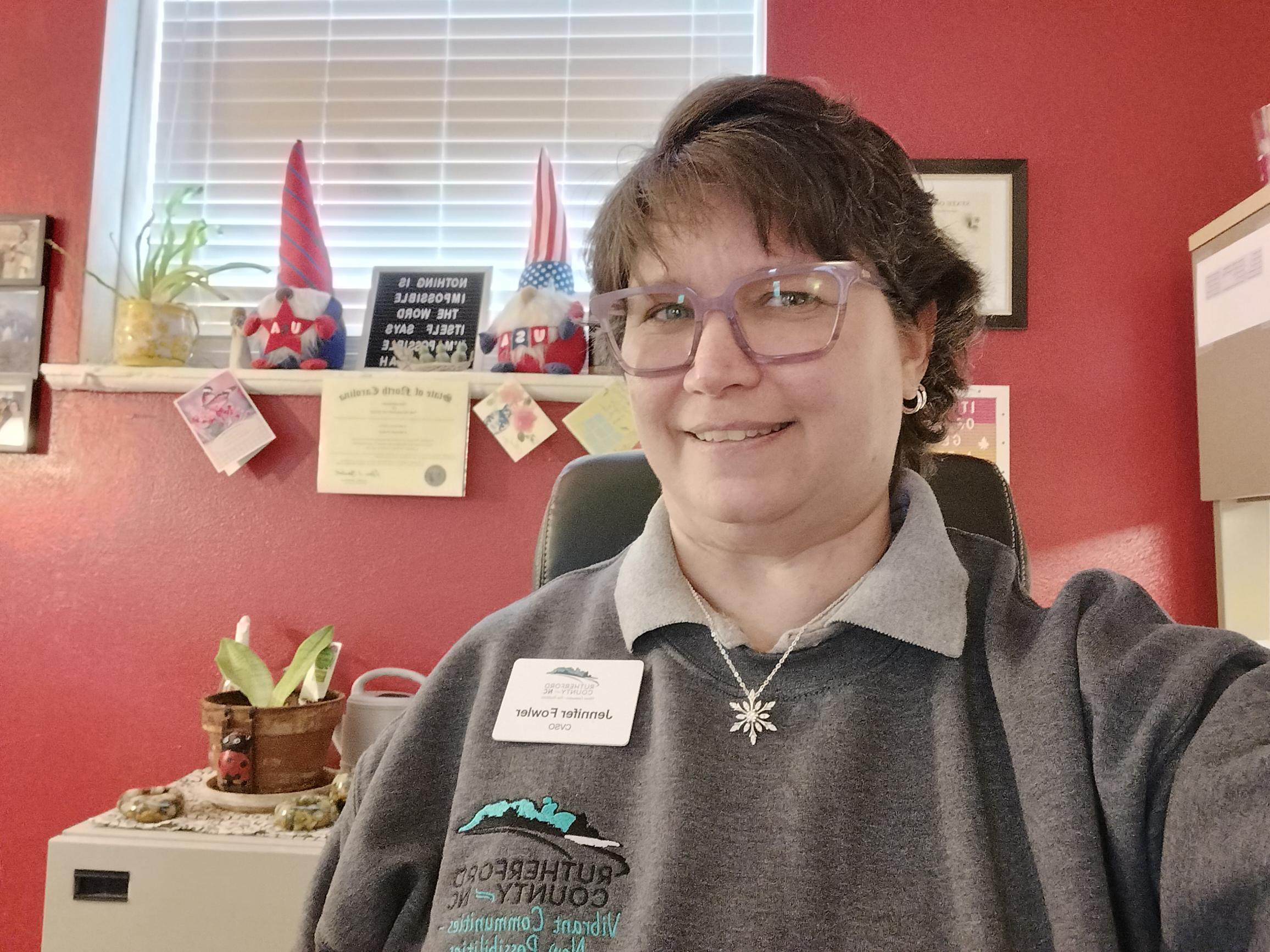 Jennifer Fowler smiles in her office wearing a name badge and gray sweatshirt